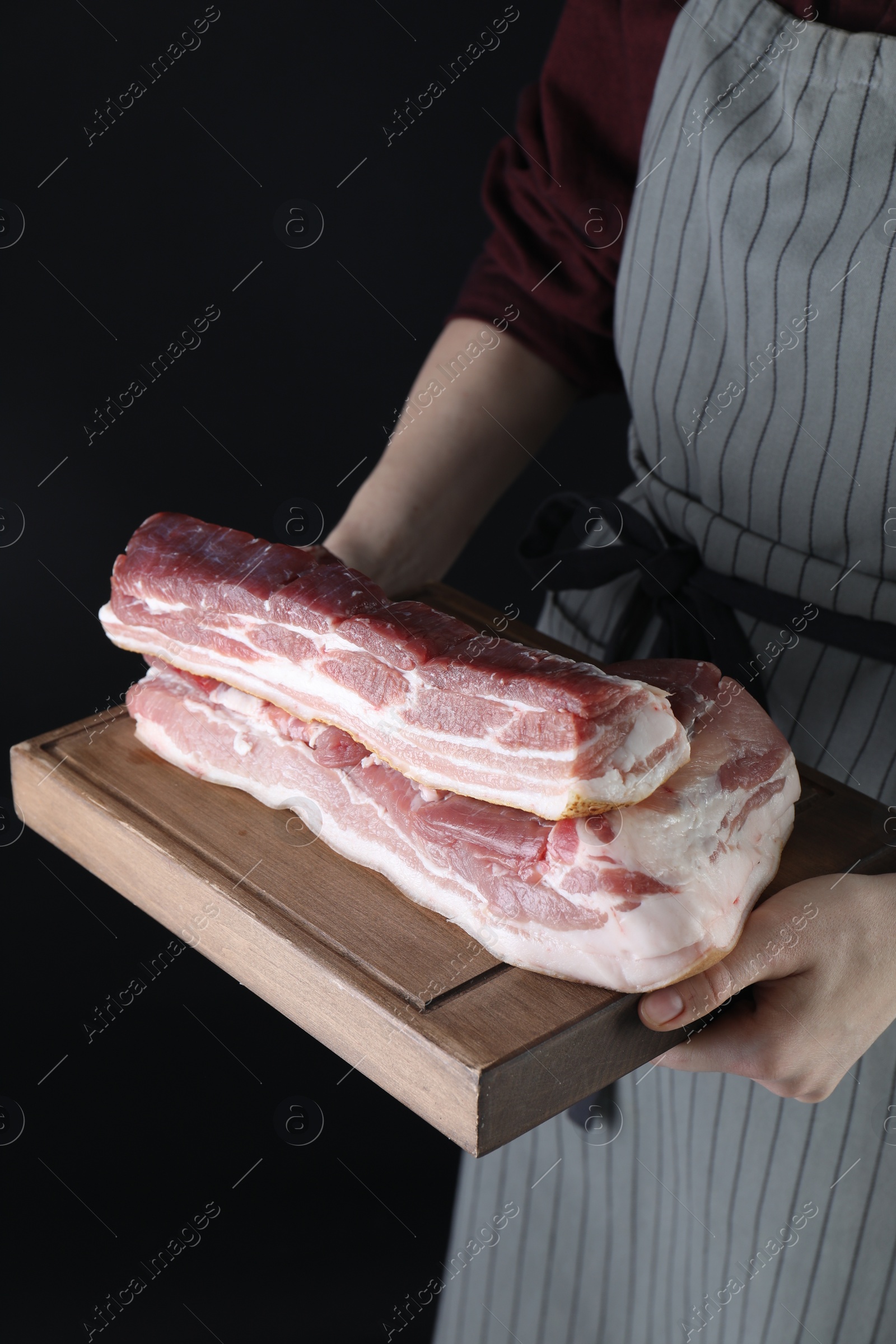 Photo of Woman holding wooden board with pieces of raw pork belly on black background, closeup