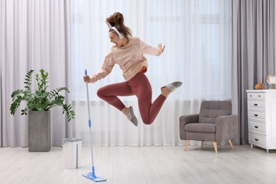 Photo of Enjoying cleaning. Woman in headphones jumping with mop indoors