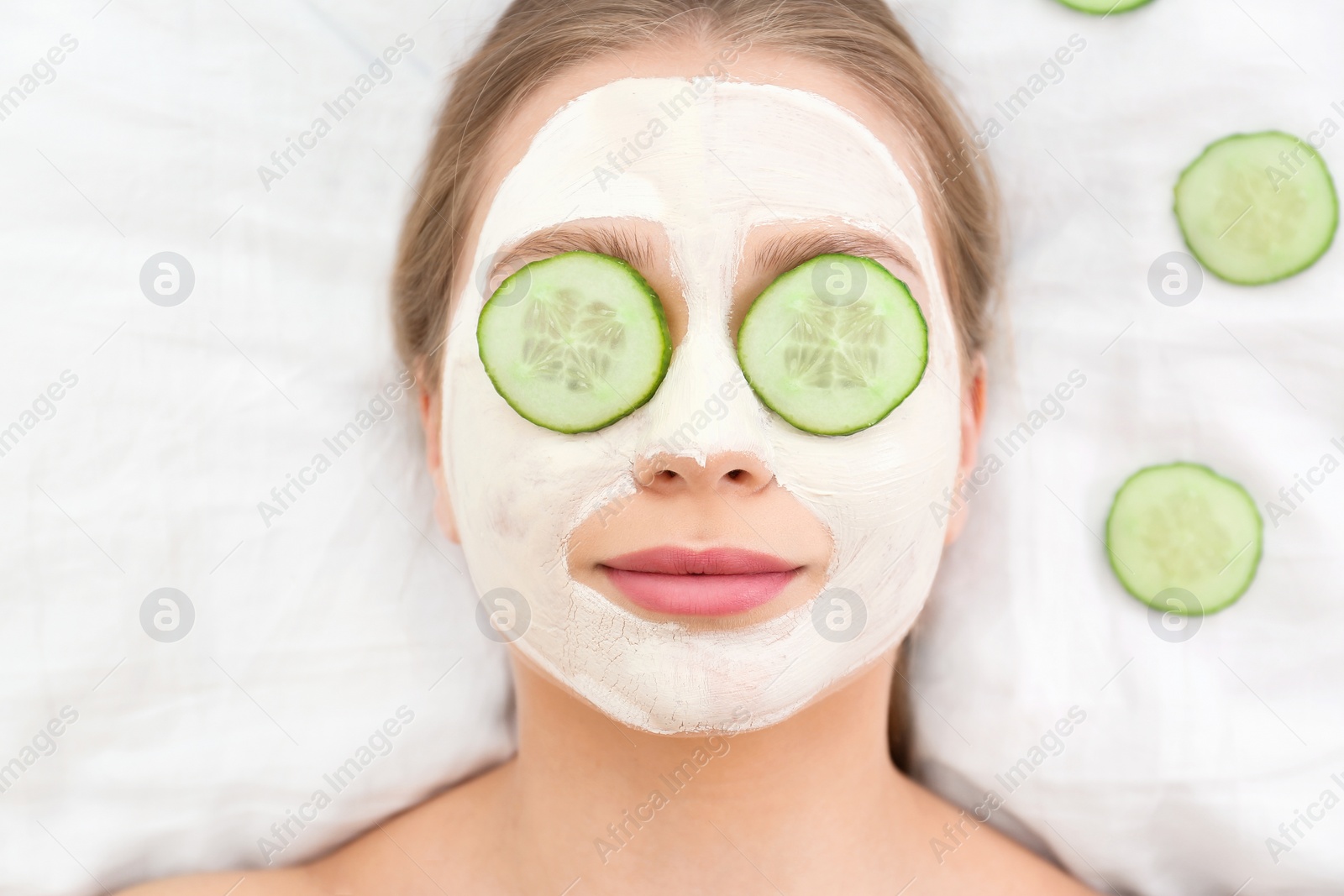 Photo of Beautiful woman with organic facial mask and cucumber slices on white fabric, above view