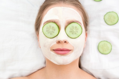 Photo of Beautiful woman with organic facial mask and cucumber slices on white fabric, above view