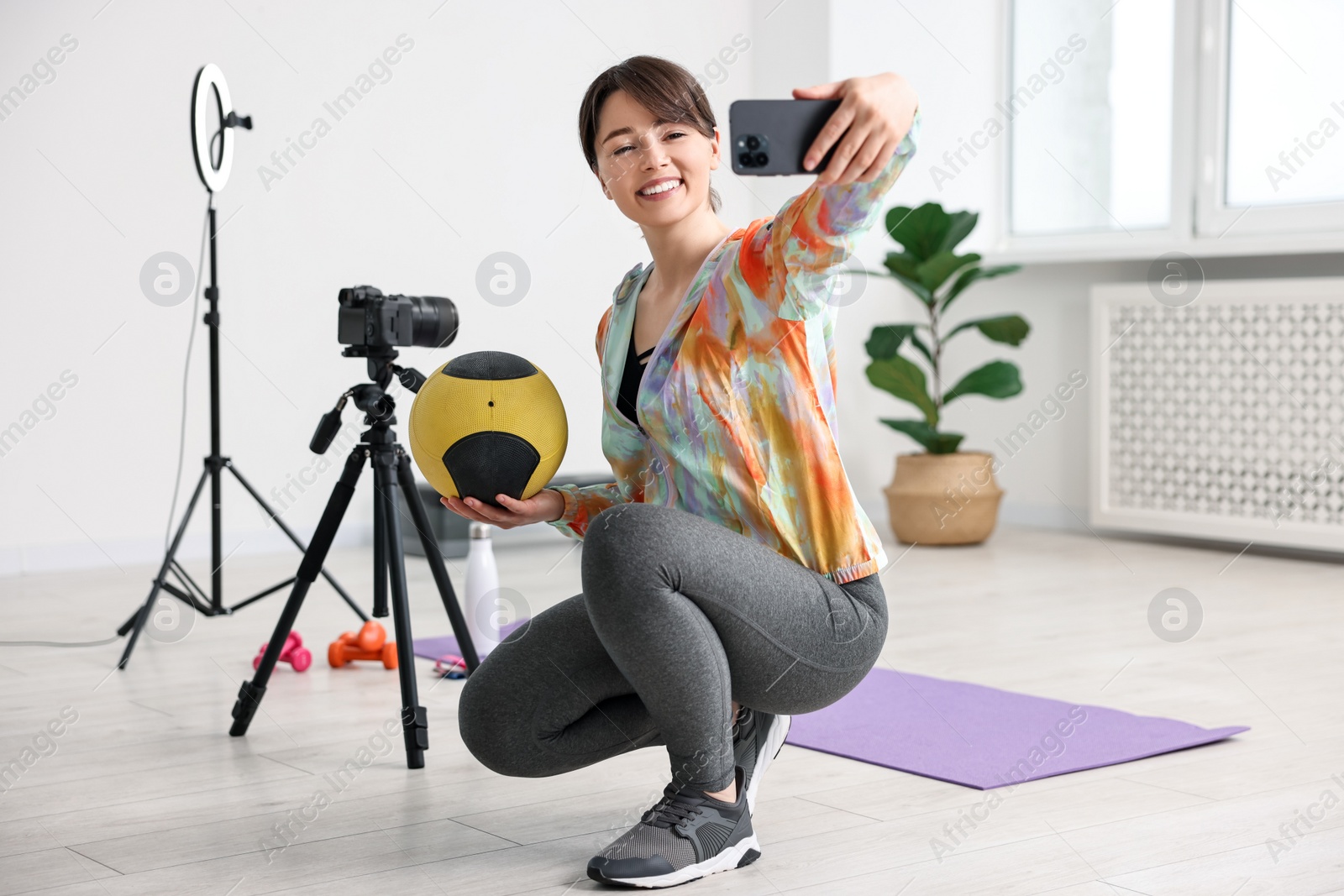 Photo of Happy sports blogger holding medicine ball while streaming online fitness lesson with smartphone at home