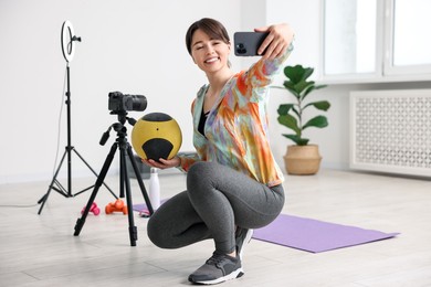 Happy sports blogger holding medicine ball while streaming online fitness lesson with smartphone at home
