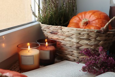 Photo of Wicker basket with beautiful heather flowers, pumpkin, burning candles and open book near window indoors