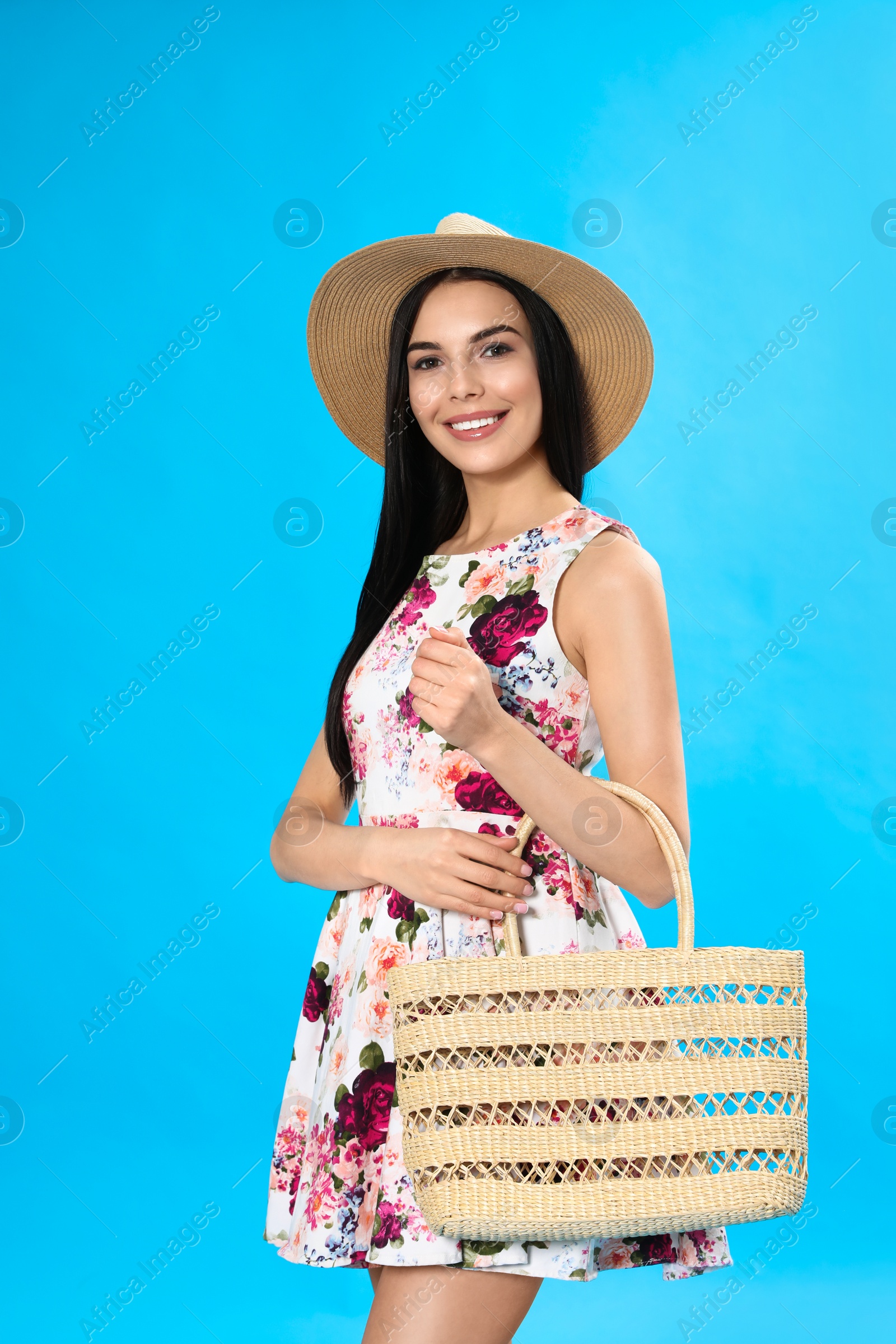 Photo of Young woman wearing floral print dress with straw bag on light blue background