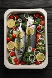 Photo of Raw fish with vegetables and lemon in baking dish on black wooden table, top view