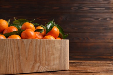 Photo of Fresh tangerines with green leaves in crate on wooden table, closeup. Space for text
