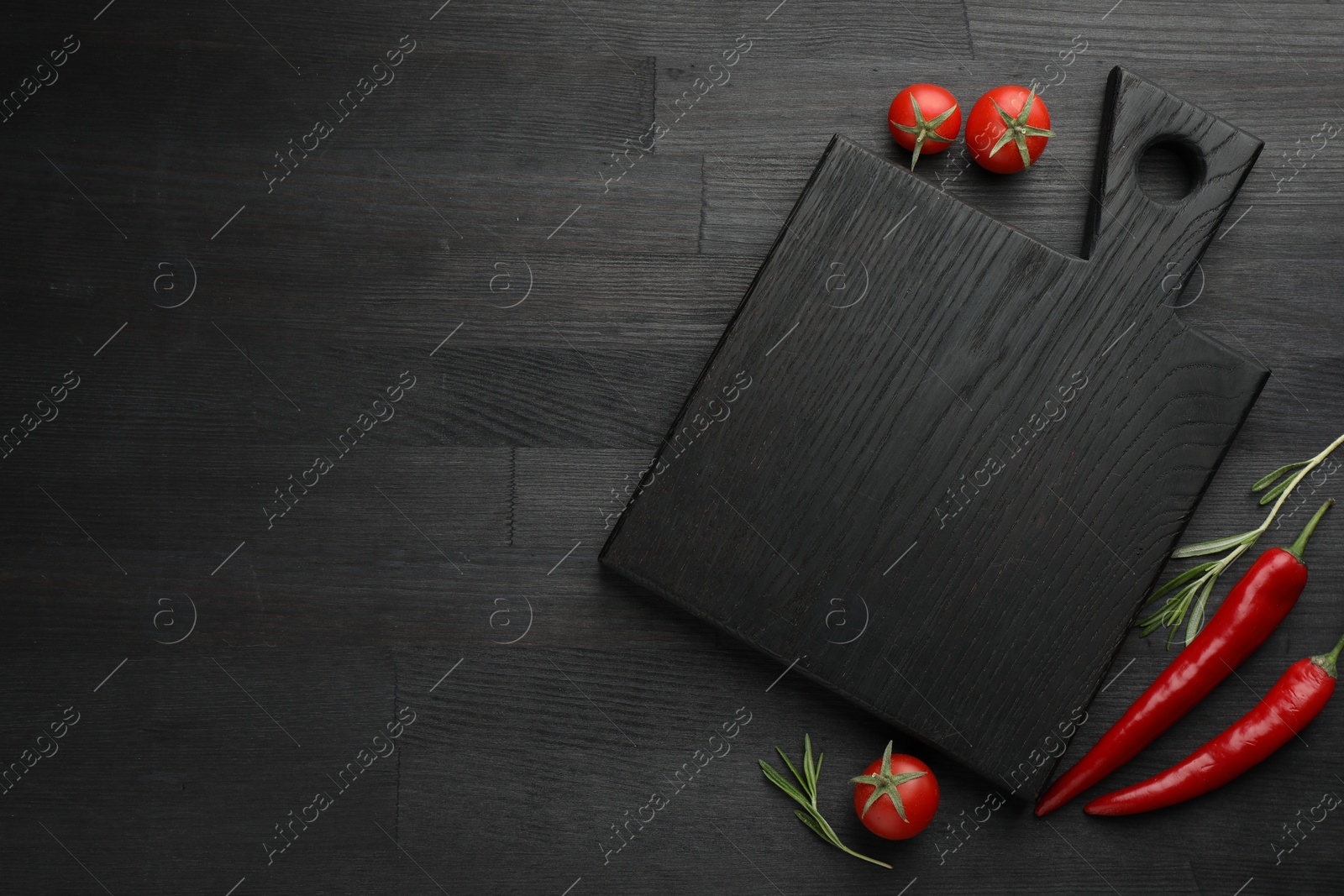 Photo of Cutting board, rosemary, chili peppers and tomatoes on black wooden table, flat lay. Space for text