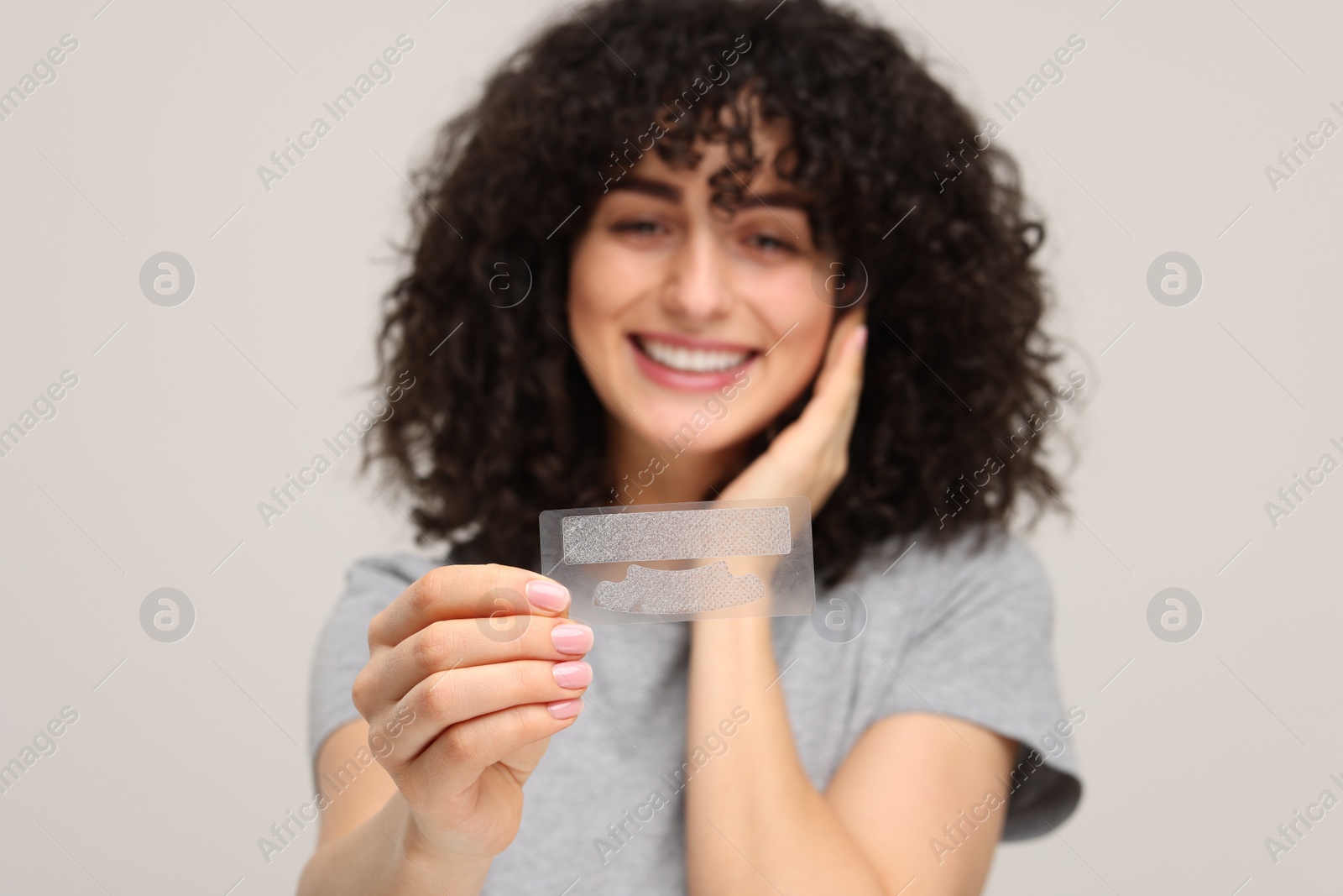 Photo of Young woman holding teeth whitening strips on light grey background, selective focus