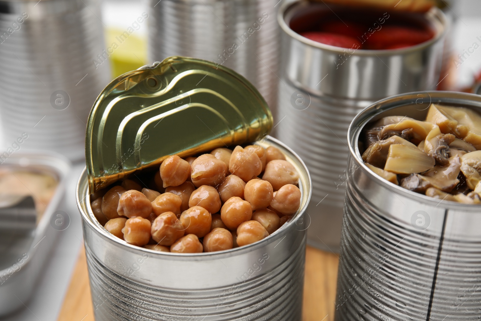 Photo of Open tin cans with different preserved products on wooden board, closeup