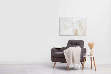 Comfortable armchair, blanket and side table near white wall indoors. Space for text