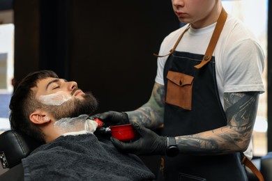 Photo of Professional hairdresser applying shaving foam onto client's beard in barbershop