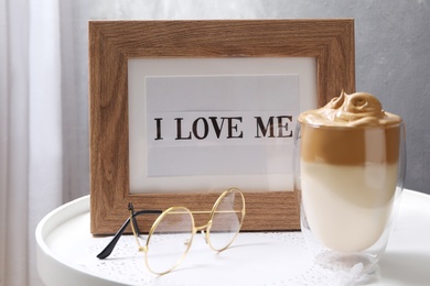 Photo of Frame with phrase I Love Me near eyeglasses and cup of coffee drink on table indoors