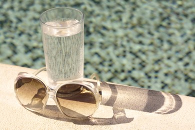 Stylish sunglasses and glass of water near outdoor swimming pool on sunny day, space for text