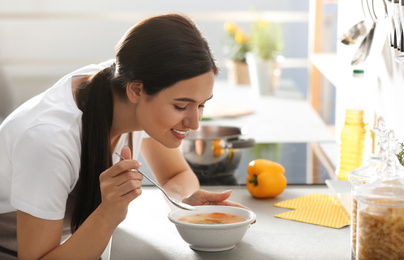 Photo of Young woman eating tasty vegetable soup at countertop in kitchen