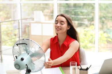 Photo of Young woman enjoying air flow from fan at workplace. Summer heat