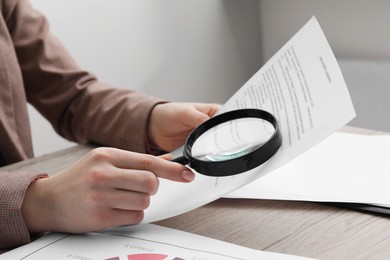 Woman looking at document through magnifier at wooden table indoors, closeup. Searching concept