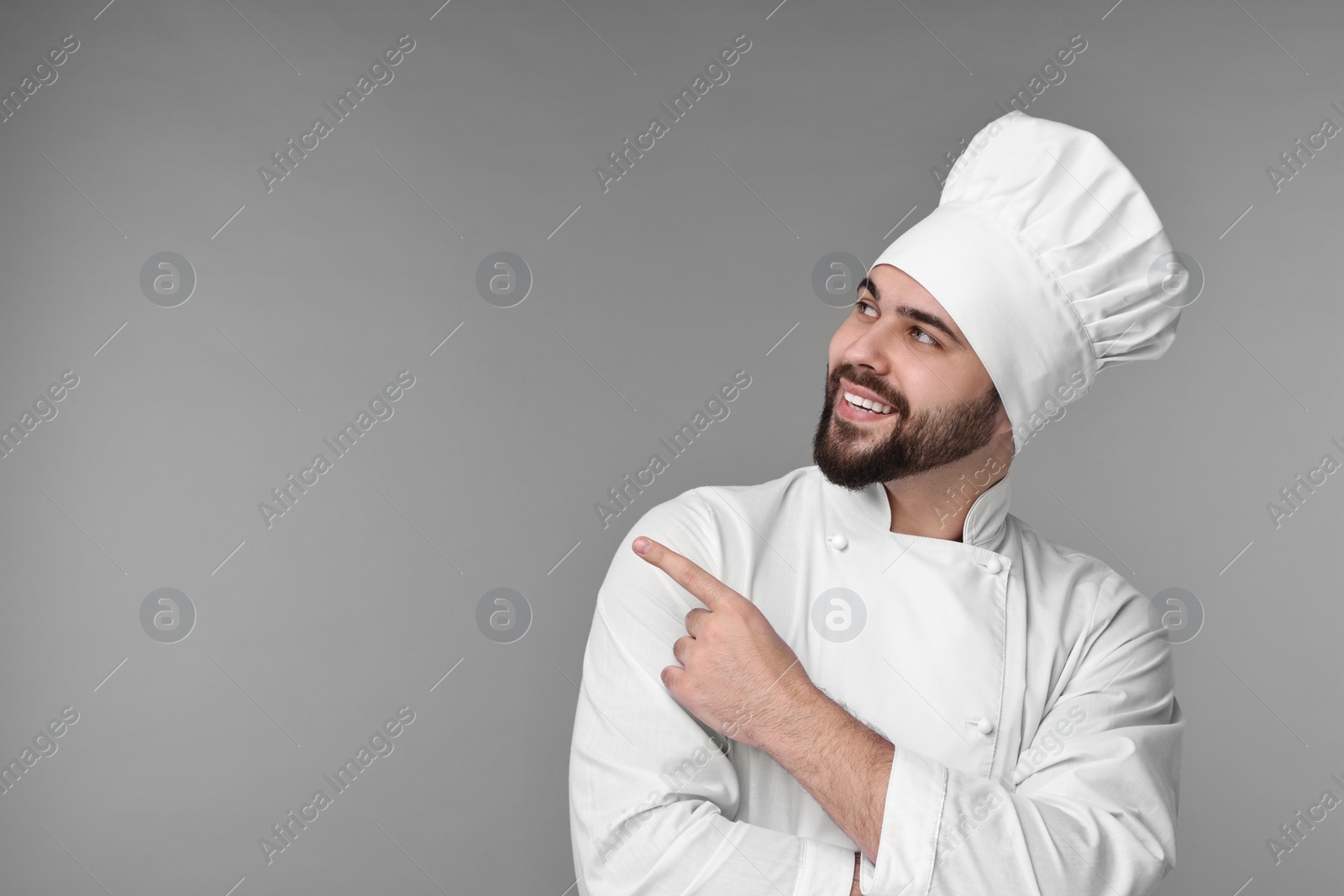 Photo of Happy young chef in uniform pointing at something on grey background. Space for text