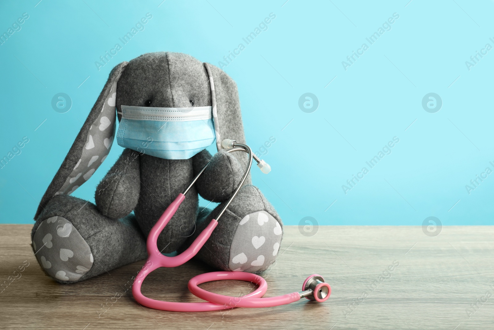 Photo of Toy bunny with protective mask and stethoscope on wooden table, space for text. Pediatrician practice