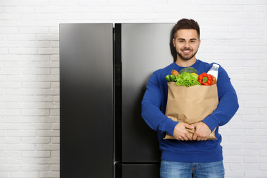 Photo of Young man with paper bag full of products near refrigerator indoors