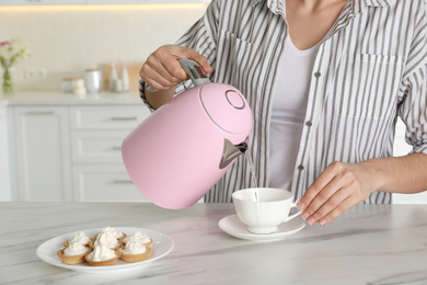 Photo of Woman pouring hot water from modern electric kettle in cup indoors, closeup