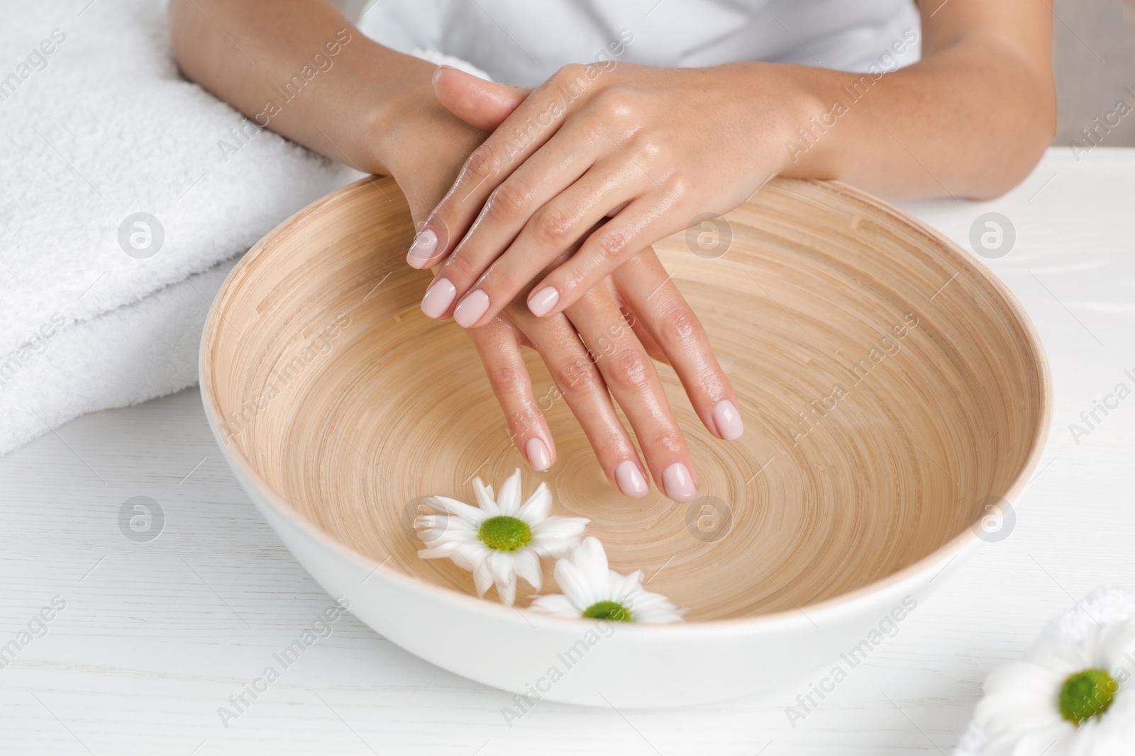 Photo of Woman soaking her hands in bowl with water and flowers on table, closeup. Spa treatment