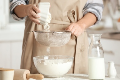 Woman sifting flour into bowl on table in kitchen