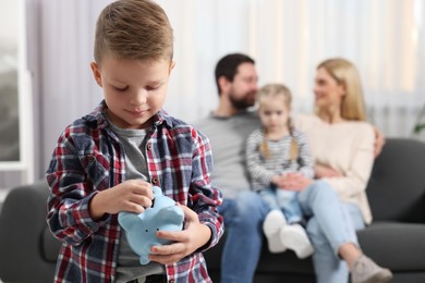 Photo of Family budget. Little boy putting coin into piggy bank while his parents and sister sitting on sofa at home, selective focus