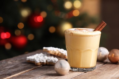 Photo of Tasty eggnog with cinnamon, cookies and baubles on wooden table against blurred festive lights. Space for text