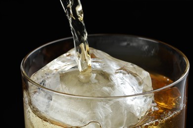 Photo of Pouring tasty whiskey into glass with ice on black background, closeup