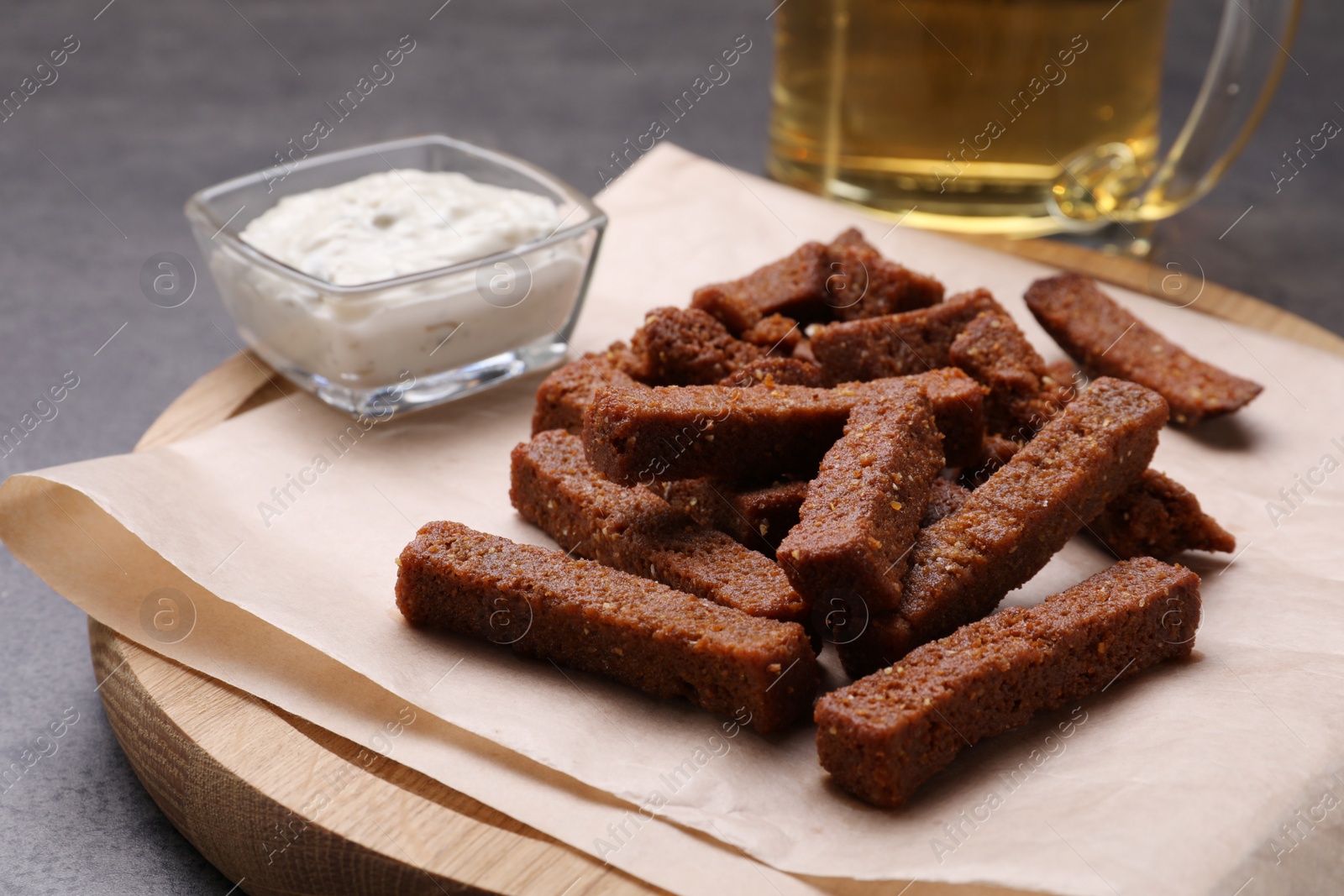 Photo of Wooden board with crispy rusks and dip sauce on grey table, closeup