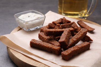 Wooden board with crispy rusks and dip sauce on grey table, closeup