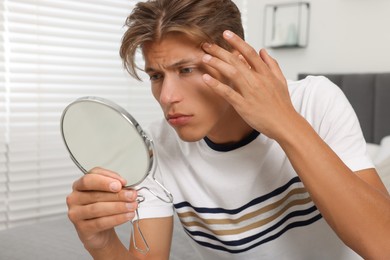 Upset young man looking at mirror and touching pimple on his face indoors. Acne problem