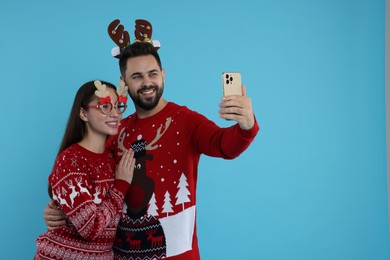 Photo of Happy young couple in Christmas sweaters, reindeer headband and party glasses taking selfie on light blue background. Space for text