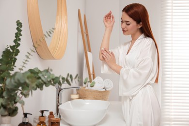 Photo of Beautiful young woman applying body cream onto elbow in bathroom, space for text
