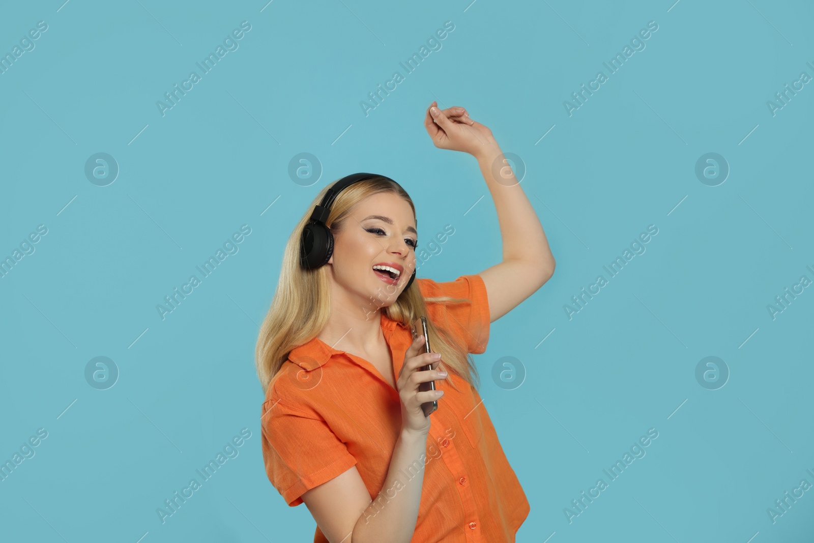Photo of Happy woman in headphones enjoying music and singing into smartphone on light blue background