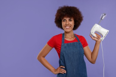 Photo of Happy young woman in apron holding mixer on purple background. Space for text