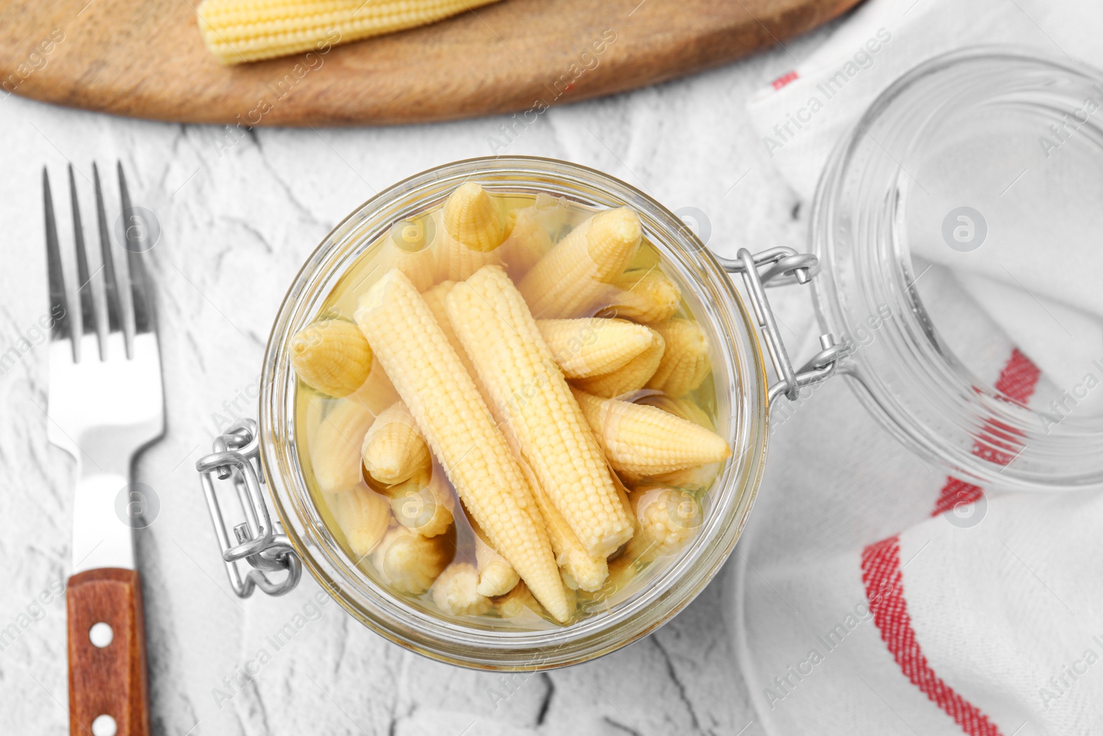 Photo of Jar of pickled baby corn and fork on white textured table, flat lay