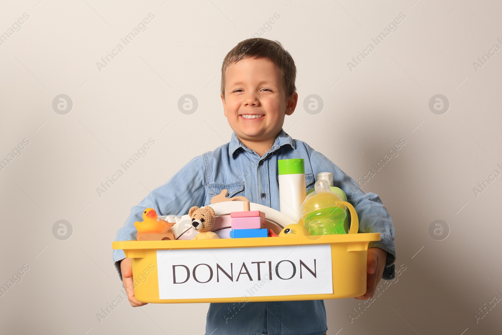Photo of Cute little boy holding donation box with goods and toys against light background