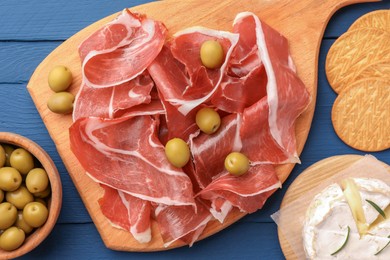 Slices of tasty cured ham, olives and cheese on blue wooden table, flat lay