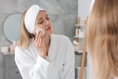 Young woman cleaning her face with cotton pad near mirror in bathroom