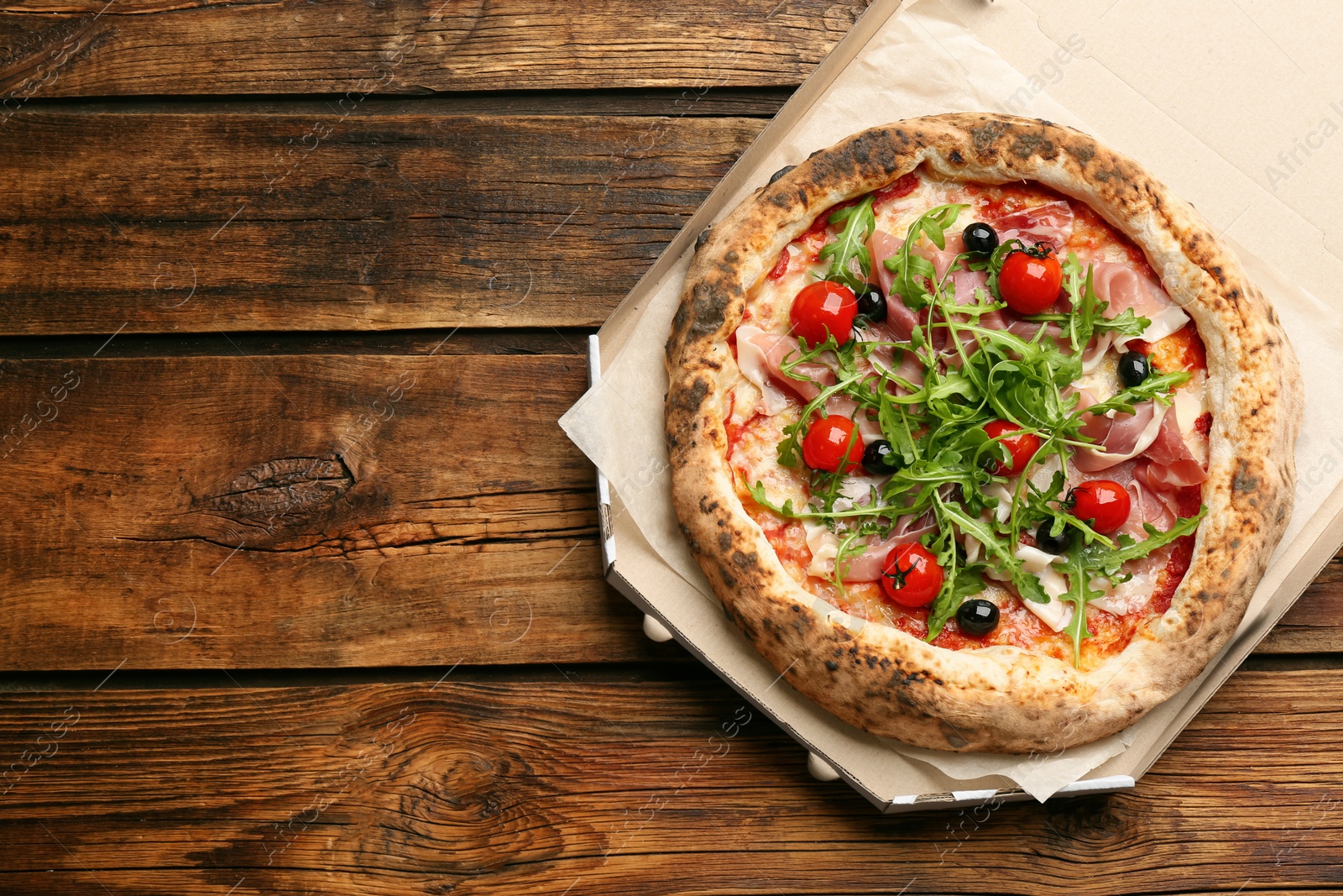 Photo of Tasty pizza with meat and arugula in cardboard box on wooden table, top view. Space for text