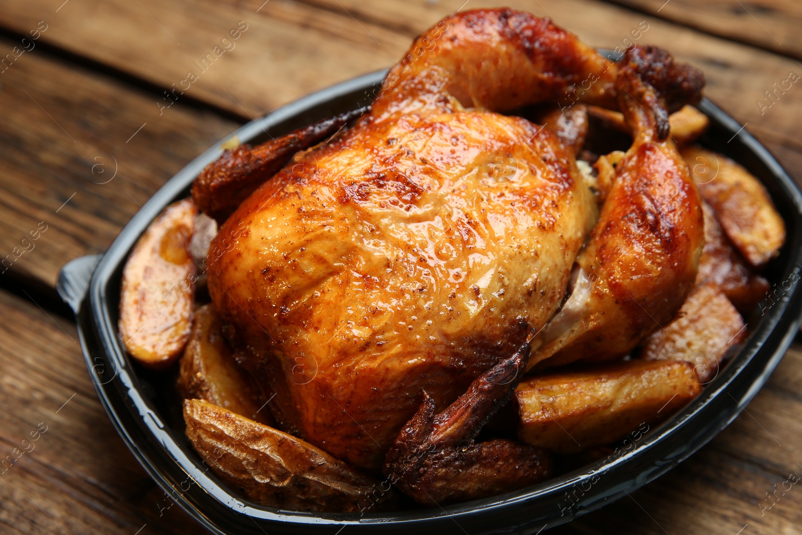 Photo of Delicious grilled whole chicken with potato in plastic container on wooden table, closeup. Food delivery service