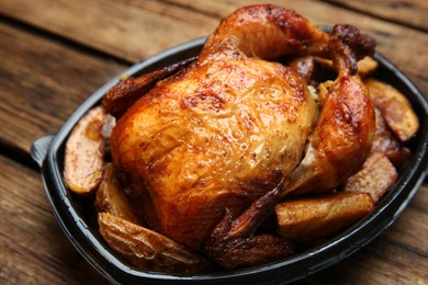 Photo of Delicious grilled whole chicken with potato in plastic container on wooden table, closeup. Food delivery service