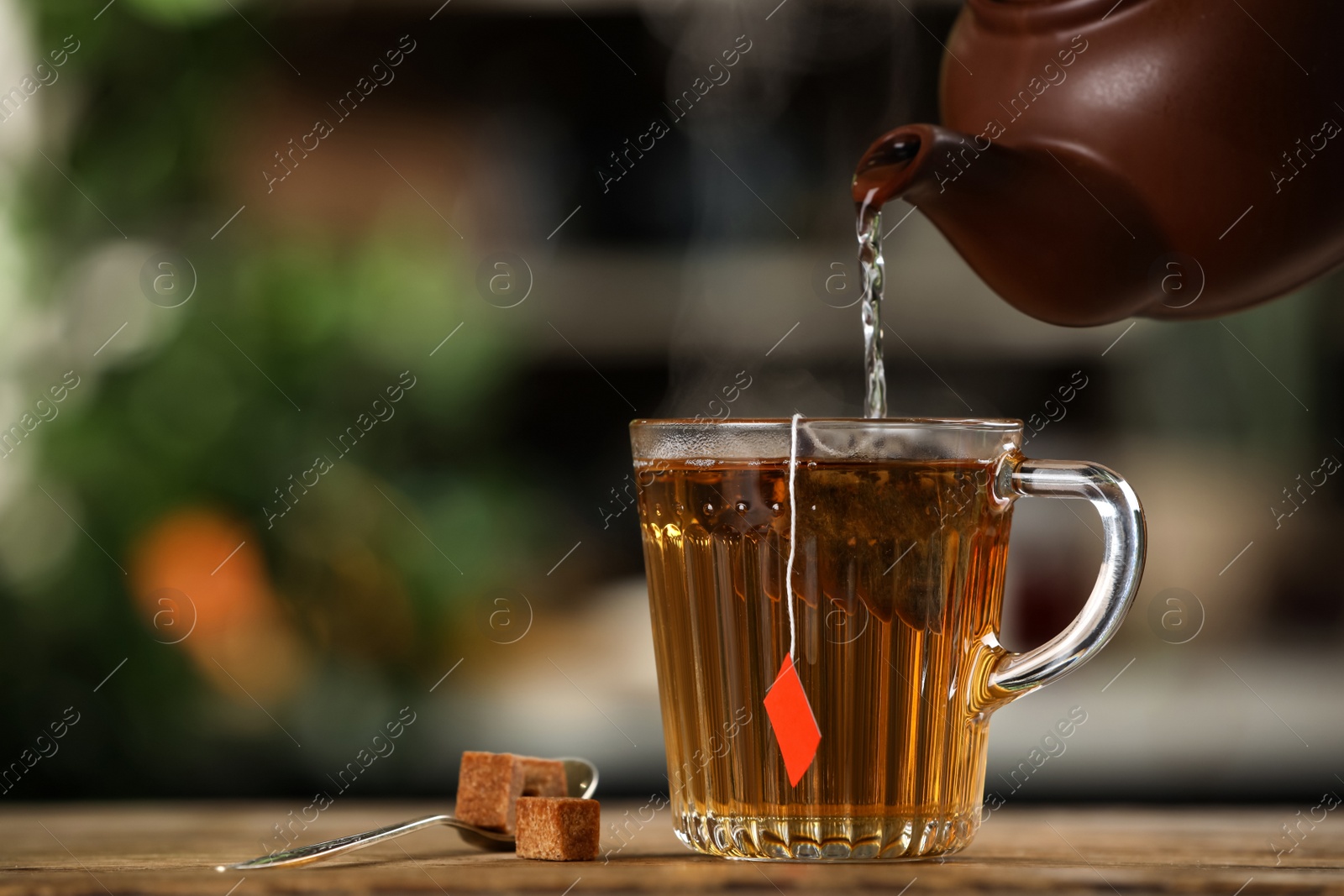 Photo of Pouring hot water into glass cup with tea bag at wooden table against blurred background, space for text