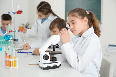 Photo of Smart girl looking through microscope and her classmates at chemistry lesson