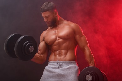 Young bodybuilder exercising with dumbbells in smoke on color background, low angle view