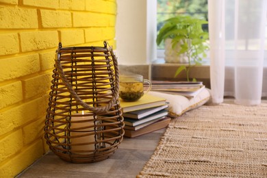 Stylish holder with burning candle and books on floor near yellow brick wall. Space for text