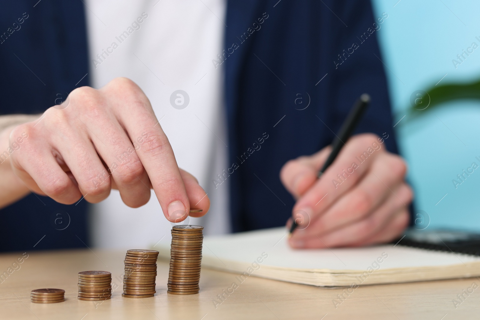 Photo of Financial savings. Man stacking coins while writing down notes at wooden table, closeup