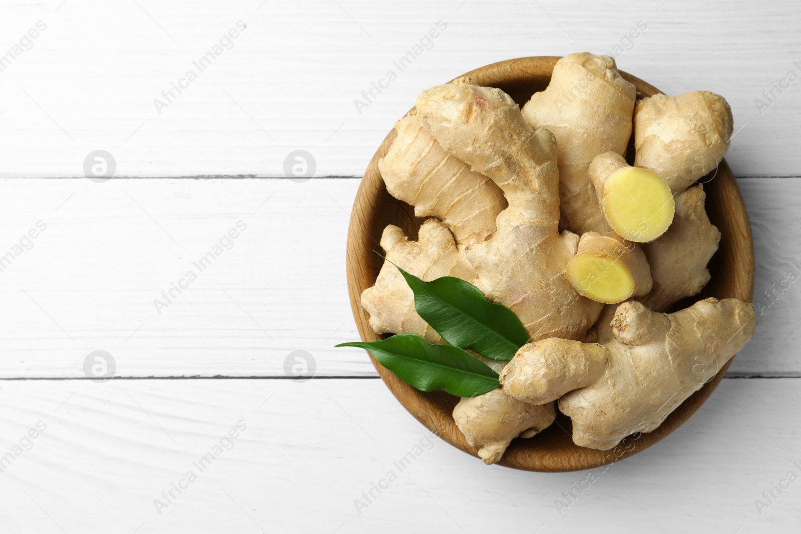 Photo of Fresh ginger with leaves in bowl on white wooden table, top view