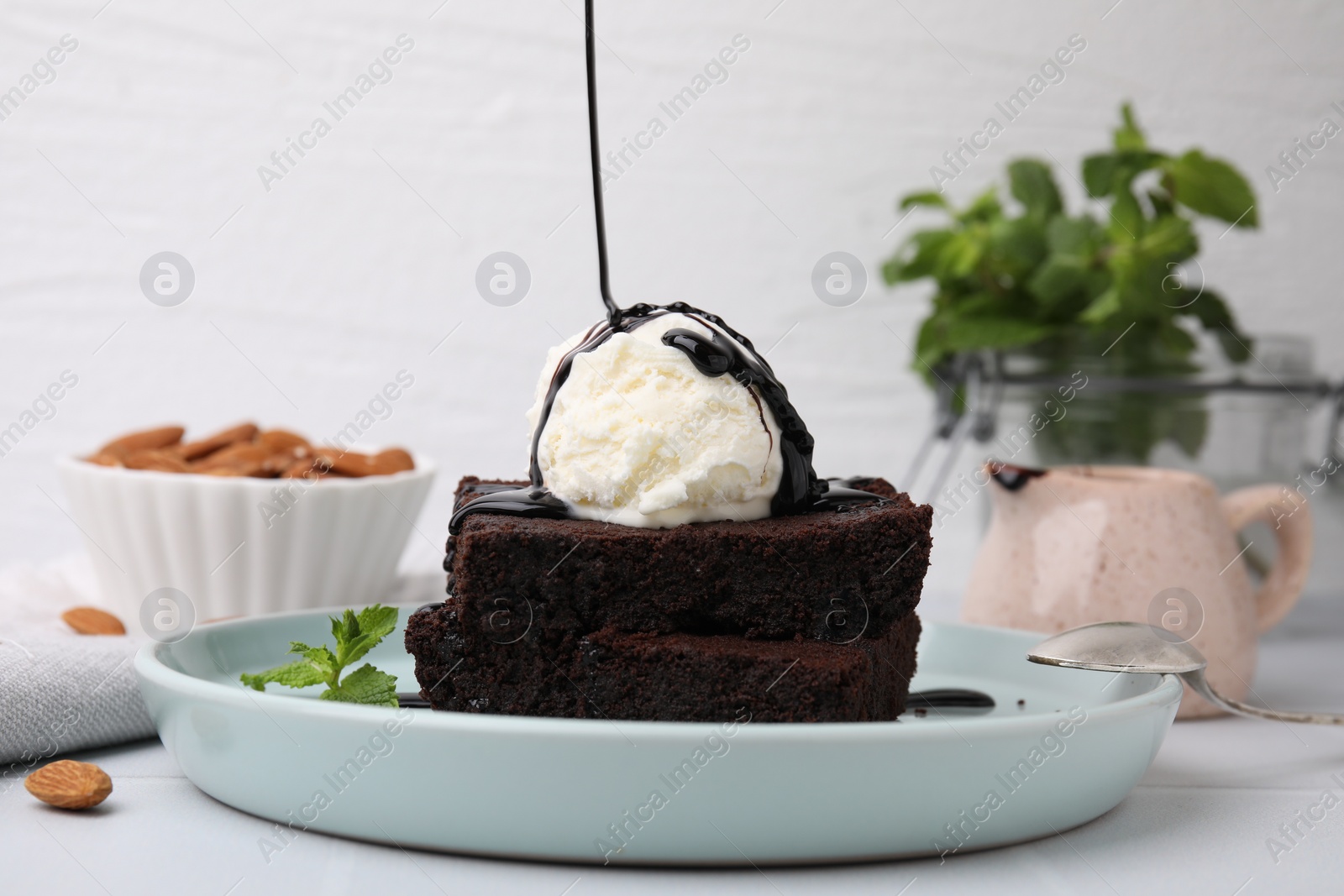 Photo of Pouring chocolate sauce onto delicious brownies with ice cream on white table, closeup